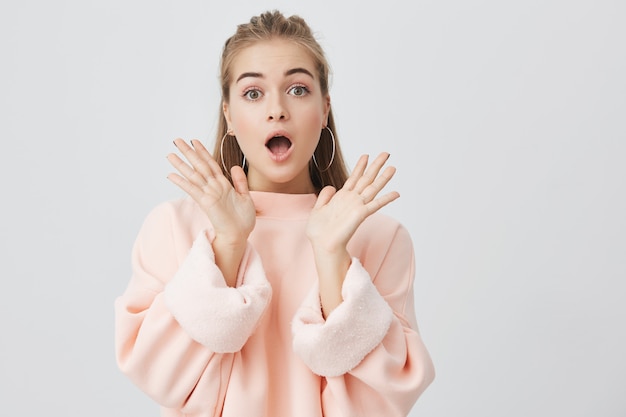 Indoor shot of excited young woman with fair hair having shocked look, exclaiming, keeping mouth wide open and hands out surprised with her unexpected victory or present.