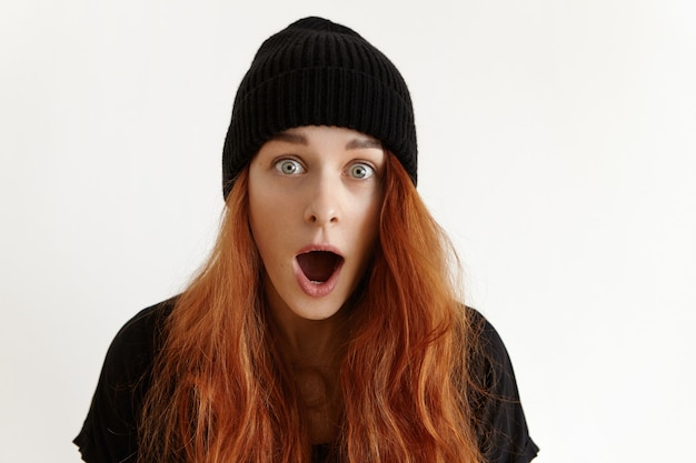 Indoor shot of fashionable astonished teenage girl with loose ginger hair wearing black hat