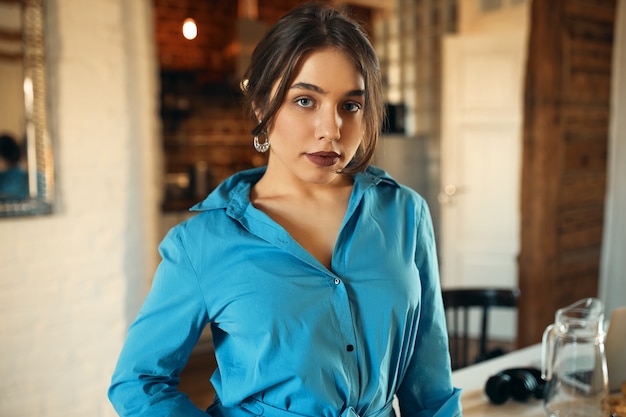 Indoor shot of fashionable stylish young Caucasian female in blue dress and earrings getting dressed going out with friends standing in living room, looking at camera with confident facial expression