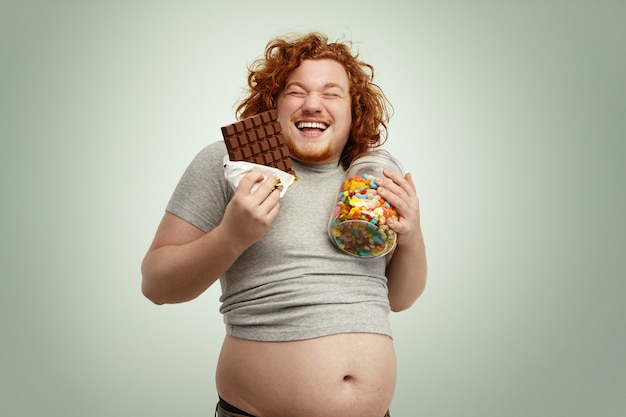 Free photo indoor shot of fat redhead male holding glass of sweets in one hand and bar of chocolate in other