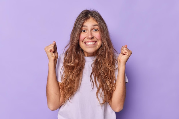 Indoor shot of glad young European woman clenches fists and bites lips awaits for important positive results dressed in casual t shirt isolated over purple wall