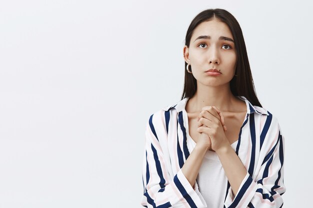 Indoor shot of gloomy cute female in striped blouse over t-shirt, clasping hands and pursing lips, frowning while gazing up with hopeful expression, waiting for results of test over gray wall