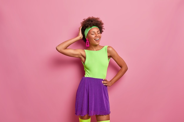 Free photo indoor shot of good looking curly haired ethnic woman with closed eyes, smiles sincerely, wears colorful t shirt and skirt