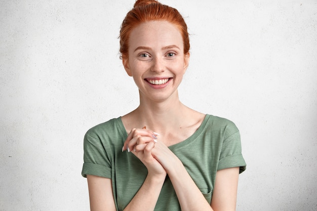 Free photo indoor shot of good looking happy female model, keeps hands together, has shining smile, wears casual clothing, being delighted