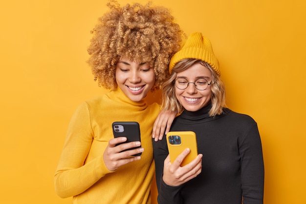 Free photo indoor shot of happy beautiful women enjoy mobile offer browse new application or website being addicted to modern technologies holds cellulars chat online isolated over vivid yellow background