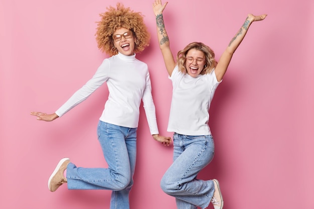 Free Photo indoor shot of happy carefree beautiful women dance and jump keep arms raise have upbeat mood cheerful expressions enjoys life isolated over pink background magnificent female friends indoor