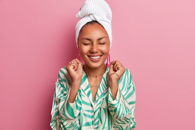 Indoor shot of happy dark skinned woman stands with eyes closed, clenches fists, rejoices good news, dressed in domestic robe, wrapped bath towel. Housewife spends weekend at home. Cozy atmosphere