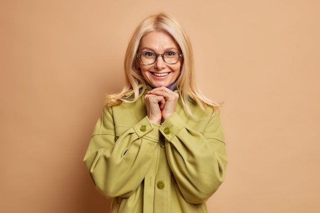 Free Photo indoor shot of happy european woman smiles toothily keeps hands under chin looks directly at camera