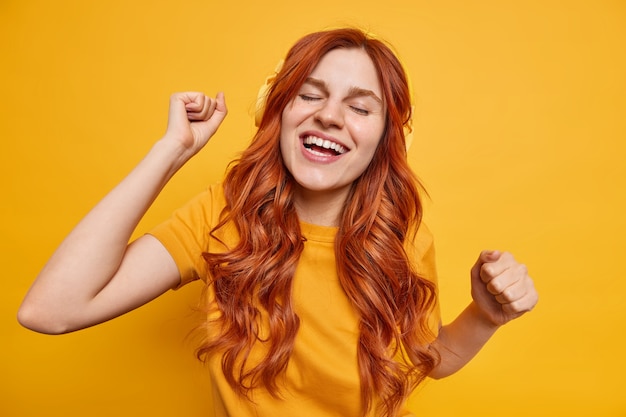 Free Photo indoor shot of happy redhead teenage girl feels relaxed shakes arms moves with rhythm of favorite music wears wireless headphones enjoys good sound smiles broadly 