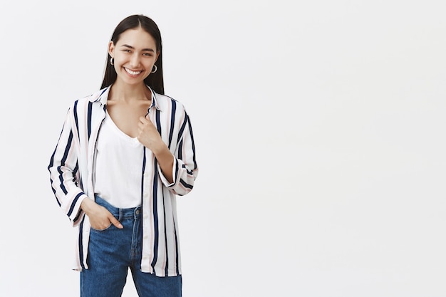 Indoor shot of happy sucessful young female entrepreneur in striped blouse and jeans, holding hand in pocket, smiling joyfully, being pleased after opening own business over gray wall