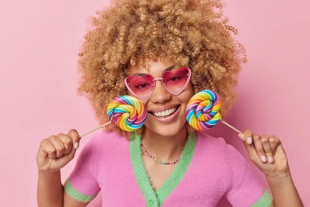 Free photo indoor shot of happy woman with curly hair holds two colorful candies near face smiles toothily wears pink sunglasses and casual t shirt isolated over pink background sweet tooth and fun concept