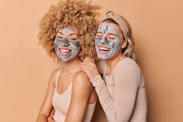 Indoor shot of happy young pretty women apply clay masks smile broadly dressed casually stand closely to each other pose against brown background Facial treatments beauty and skin care concept