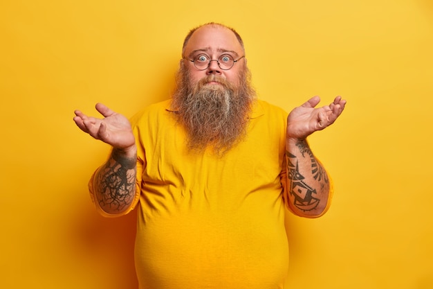 Free Photo indoor shot of hesitant bearded man with excess weight shrugs shoulders and stands unaware, has thick beard, big beer belly, dressed in yellow t shirt, round spectacles, faces difficult choice.