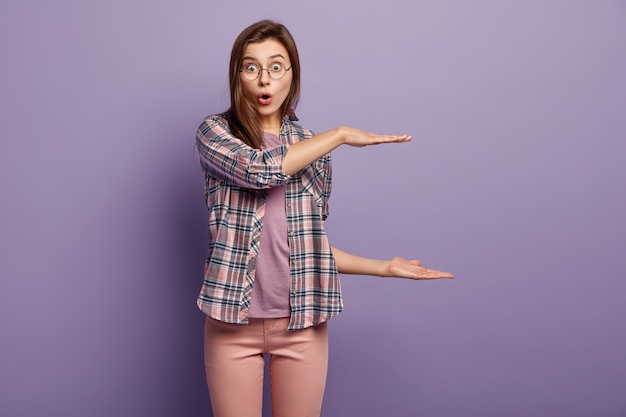 Indoor shot of impressed stupefied woman shapes big object with both hands, surprised facial expression