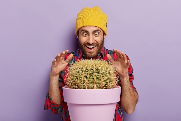 Free photo indoor shot of joyful male florist raises palms over prickly cactus in pot, has surprised happy look, dressed in stylish hat and plait shirt, isolated over purple background. man with indoor plant