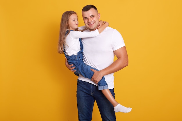 Indoor shot of loving daddy holding at little adorable daughter feeling love isolated over yellow