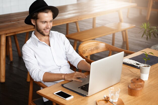 Indoor shot of male blogger typing on keyboard of laptop, using free wi-fi at modern cafe while working on his new post on social networks, looking at screen with happy and inspired face expression