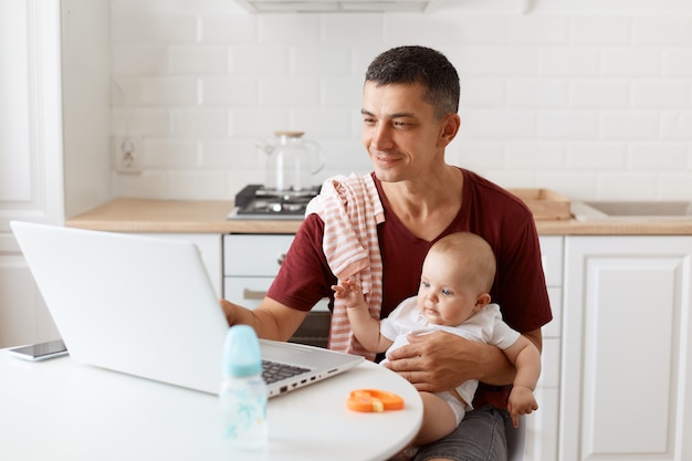 Indoor shot of man wearing burgundy casual t shirt with towel on his shoulder, looking after baby and working online from home, looking smiling at notebook screen.