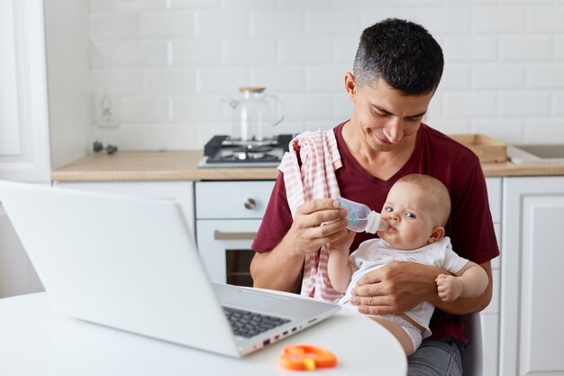 Indoor shot of man wearing maroon casual t shirt holding baby bottle, little daughter or son drinking water with father's hands, family posing at table in kitchen.