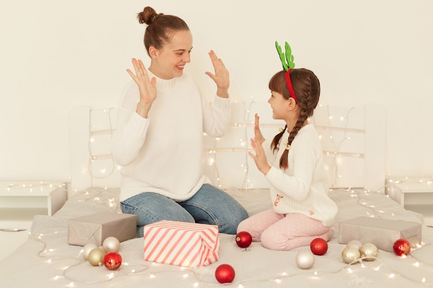 Indoor shot of optimistic mother and daughter wearing casual style white sweaters sitting on bed, giving five to each other, celebrating Christmas, happy new year.