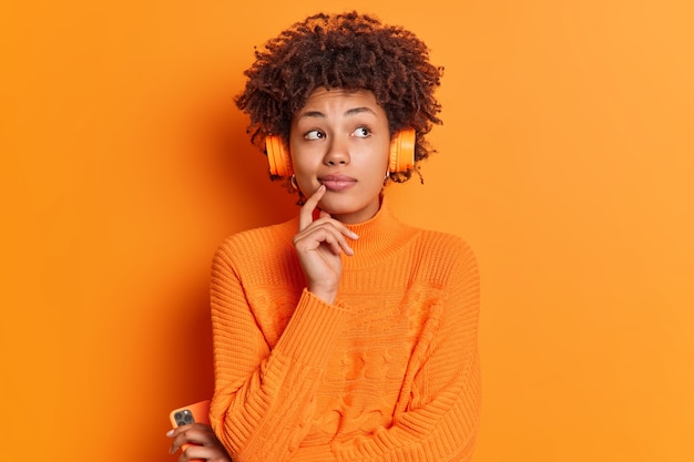 Free photo indoor shot of pensive afro american woman concentrated above keeps finger on chin considers something in mind while listening calm melody in stereo headphones