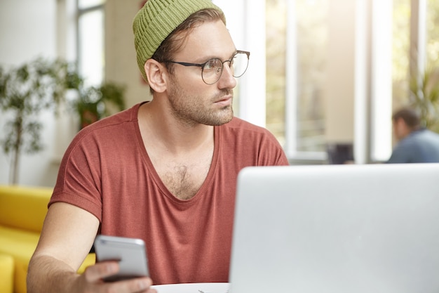 Free photo indoor shot of pensive male writer works on writing new book or chapter, holds smart phone in hand