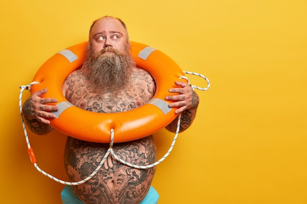 Free photo indoor shot of pensive overweight man looks away, ready for recreation, swimming in sea with lifebuoy, has naked body, isolated on yellow wall, blank space aside. safety equipment, rescue