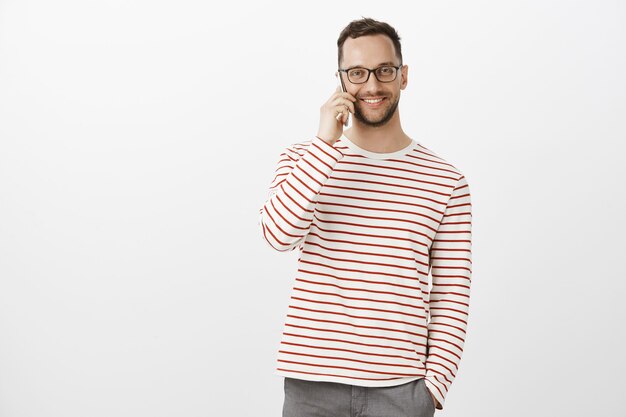 Indoor shot of pleased friendly adult office worker in striped pullover and glasses, talking on smartphone casually