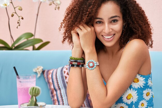 Indoor shot of pleased young beautiful female has Afro hairstyle, dressed in summer clothing, poses on blue sofa with cocktail in cozy cafe with orchid