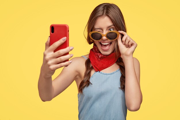 Indoor shot of pleased young brunette with sunglasses posing against the yellow wall