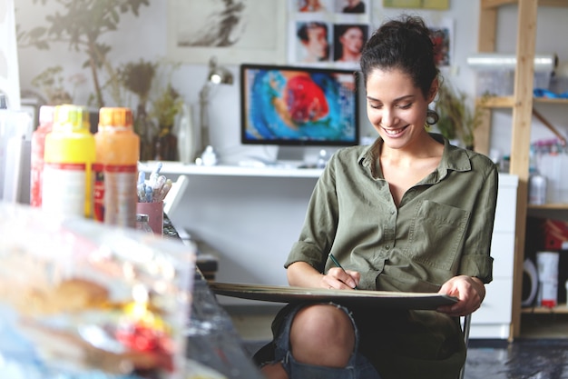 Indoor shot of positive happy young Caucasian female artist smiling broadly while working on painting or sketches at workshop; painting stuff