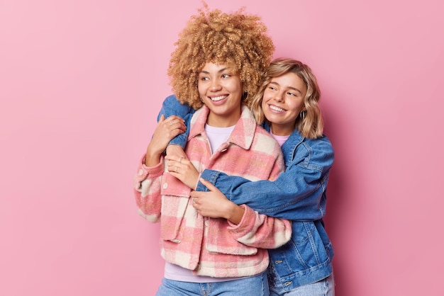 Free Photo indoor shot of positive young female friend embrace and have good relationships dressed in fashionable clothes look gladfully into distance isolated over pink background friendship concept