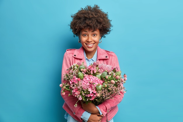 Indoor shot of positive young woman embraces big bunch of flowers smiles pleasantly dressed in pink jacket isolated over blue wall