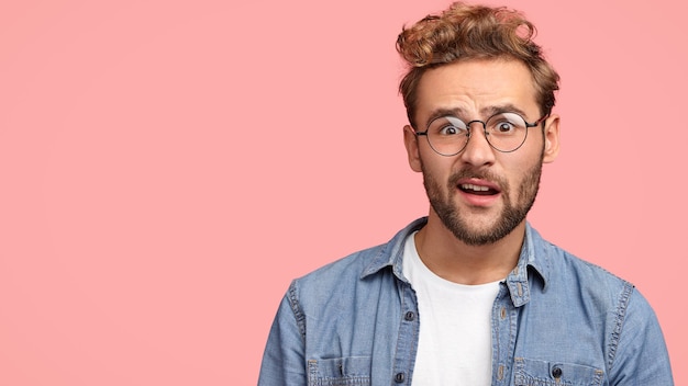 Free Photo indoor shot of puzzled frustrated male frowns face and looks with bewilderment, reacts on sudden message, dressed in fashionable clothes, isolated over pink wall