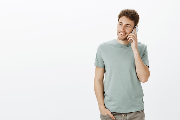 Indoor shot of relaxed charming european man with fair hair, holding smartphone near ear and gazing left with broad smile, talking casually with sister