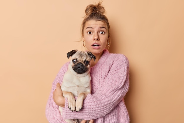 Free Photo indoor shot of scared european woman with combed hair holds pug dog carries favorite puppy wears knitted sweater isolated over brown background loves pet very much. domestic animals concept.