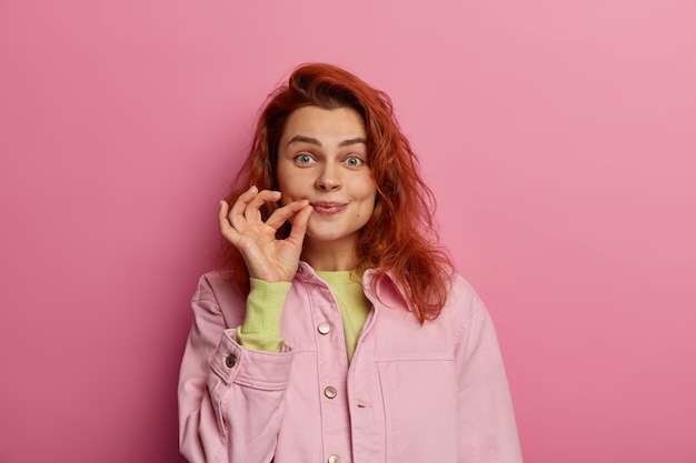 Indoor shot of secret redhead young woman zips her mouth shut, promises to keep secret, has gossip safe, holds fingers near cheek