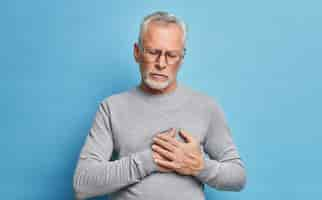 Free photo indoor shot of senior bearded man has heart attack suffers from painful feelings needs painkillers presses hands to chest being unhealthy wears spectacles and grey casual jumper isolated on blue wall