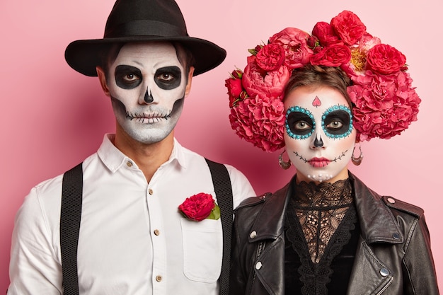 Indoor shot of serious romantic couple pose before Halloween event, wear flower wreath and hat on heads, traditional scary costumes, look directly at camera, have zombie makeup in mexican style