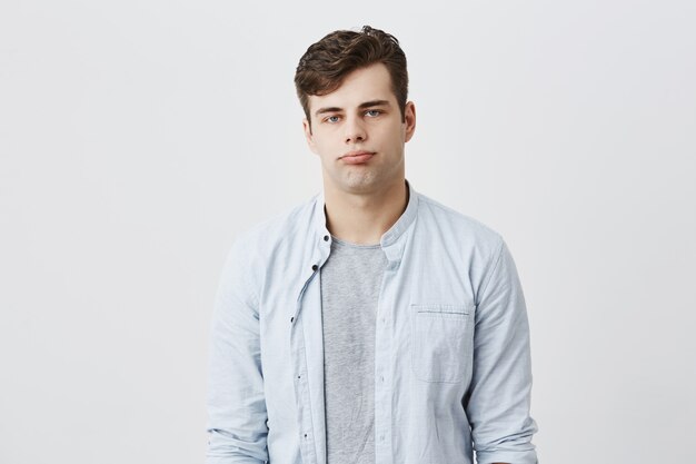 Indoor shot of serious young european male frowns face and pouts lips, being offended by friend who jokes at him, expresses dissatisfaction and discontent. People and facial expressions concept