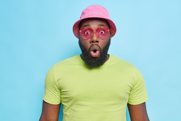 Indoor shot of shocked Afro American man with thick beard keeps mouth opened and looks scared gasps and stares concerned hears surprising news dressed casually isolated over blue wall