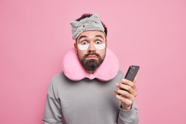 Indoor shot of shocked man checks email box via smartphone stares surprised, wears sleepmask beauty patches to reduce puffiness under eyes after sleeping 