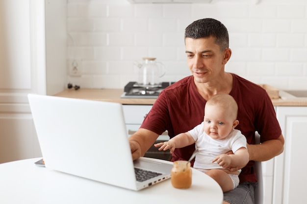 Indoor shot of smiling handsome freelancer male wearing burgundy t shirt, posing in white kitchen, sitting in front of laptop with baby in hands, typing on notebook.