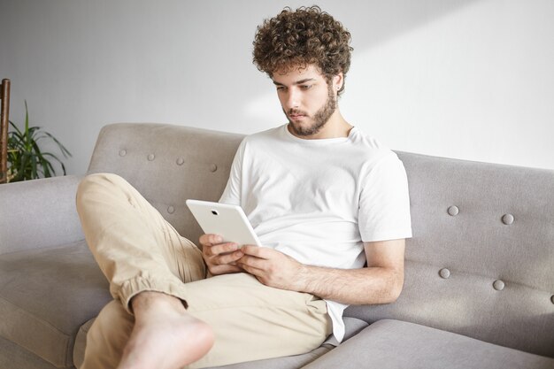 Indoor shot of stylish young Caucasian businessman in casual wear reading business news or checking email on touch pad, sitting on sofa before work. People, job, technology and occupation concept