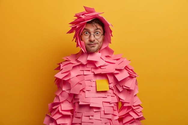 Free photo indoor shot of surprised funny man looks through transparent glasses, wears paper costume has direct gaze  isolated over yellow wall. wondered guy dressed in outfit made of sticky notes