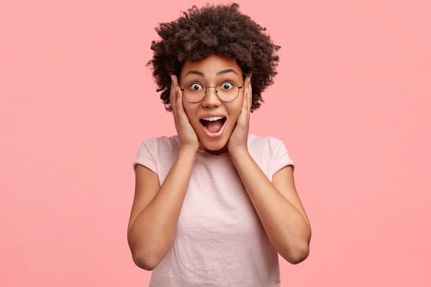 Indoor shot of surprised glad female with appealing appearance, black curly hair and dark skin, stares with eyes full of disbelief, keeps hands on cheeks, recieves something unexpected and expensive