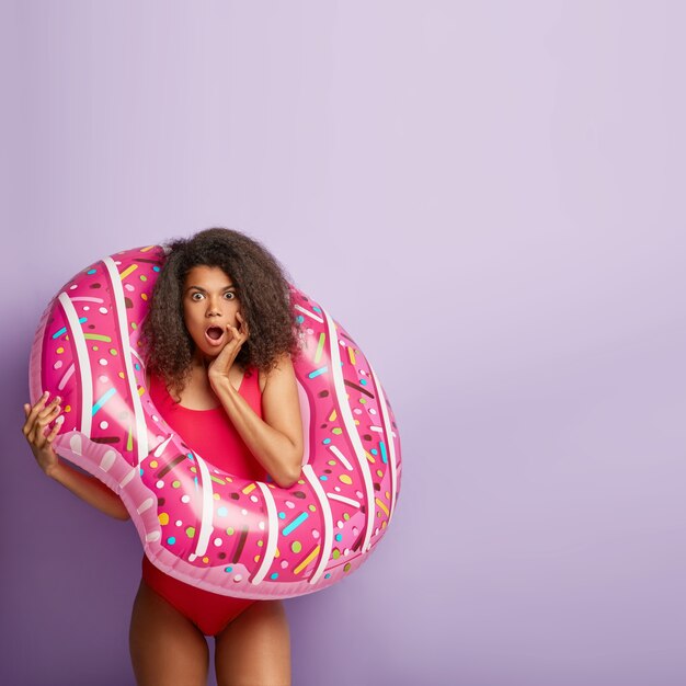 Indoor shot of surprised woman with Afro hair, poses with inflated swimring