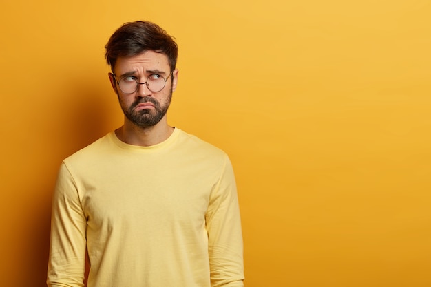 Free photo indoor shot of upset bearded young man looks with miserable expression aside, thinks about something, has sad unhappy look, wears optical glasses and casual jumper, poses indoor over yellow wall