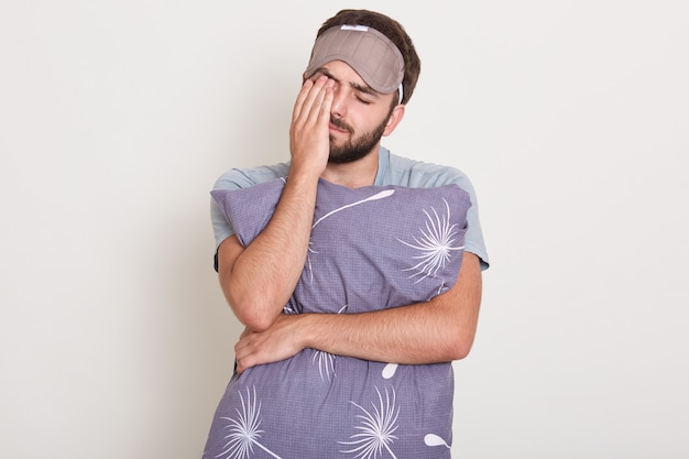 Indoor shot of young attractive man waking in morning, wants to sleep, handsome guy stretching and yawning, holding his pillos