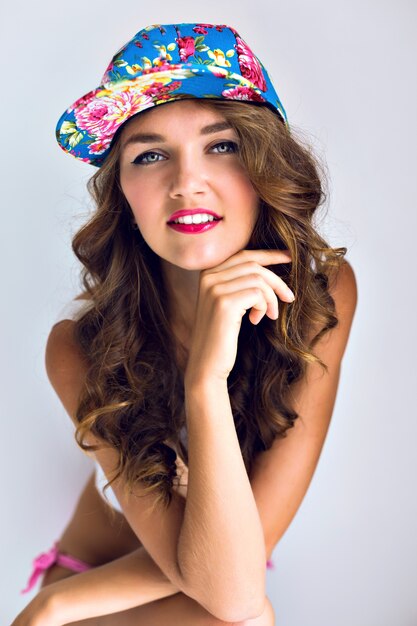 Indoor summer portrait of young sensual sexy sports tanned woman posing against a white wall in a floral swag cap and have fun alone, bright makeup , curled hairs.